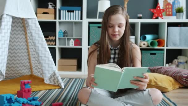 Chica alegre lectura libro disfrutando de la historia sonriendo sentado en el suelo en la habitación acogedora — Vídeo de stock
