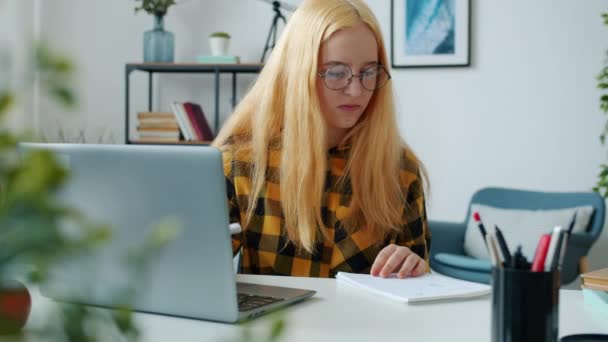Анонімна дитина вивчає онлайн з ноутбуком і робить нотатки за столом у вітальні — стокове відео
