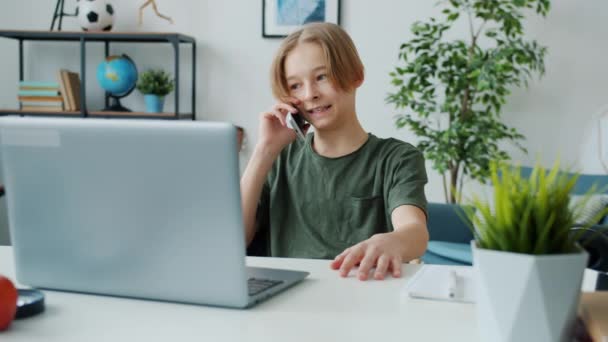 Wesoły dzieciak rozmawiający przez telefon komórkowy siedzący przy biurku komputera w domu — Wideo stockowe