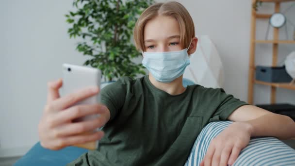 Мальчик в медицинской маске делает селфи со смартфоном дома во время пандемии — стоковое видео