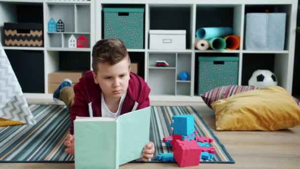 Sério menino lendo livro deitado no chão, em seguida, sorrindo apreciando história interessante — Vídeo de Stock