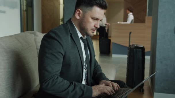 A szálloda halljában dolgozó laptopot használó üzletember, míg a női vendég a háttérben jelentkezik be — Stock videók