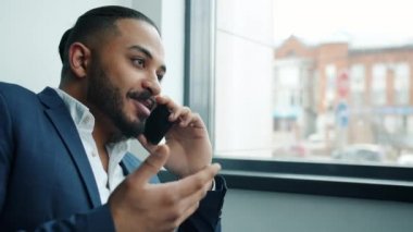 Takım elbiseli kendine güvenen bir ırk adamı cep telefonuyla konuşuyor. Pencerenin yanında iş konuşuyor.