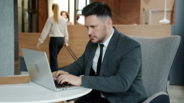 Poważny biznesmen wpisując za pomocą laptopa w holu hotelu, podczas gdy kobieta check-in w recepcji w tle — Wideo stockowe