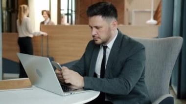 Takım elbiseli neşeli adam akıllı telefon mesajları kullanıyor. Gülümsüyor, otel lobisinde oturuyor.