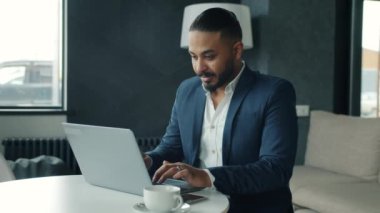 Takım elbiseli, laptopla çalışan, modern ofiste tek başına oturan melez bir adam.