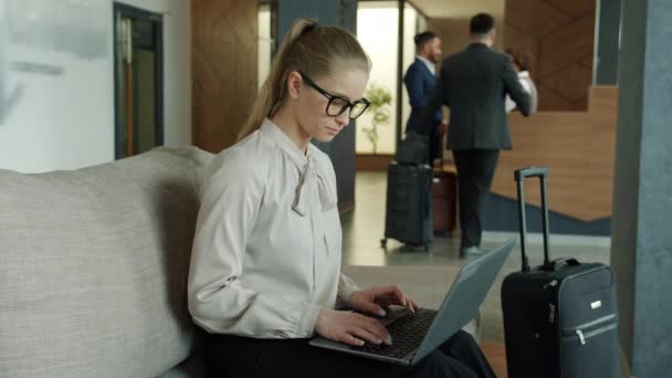 Fiatal nő dolgozik laptop a hotel halljában, míg a férfiak bejelentkeznek a recepción — Stock videók