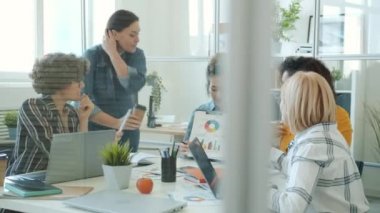 Neşeli bayan meslektaşlar ofiste takım olarak çalışırken dizüstü bilgisayar ve tablet kullanarak sohbet ediyorlar.