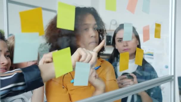 Equipo creativo multiétnico discutiendo la escritura de negocios en notas adhesivas coloridas en tablero de cristal — Vídeo de stock