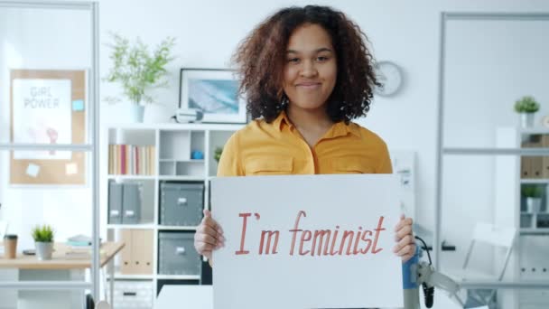 Retrato de la alegre chica afroamericana sosteniendo pancarta feminista sonriendo de pie en el interior sola — Vídeos de Stock