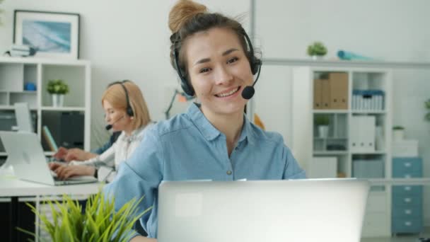 Slow motion porträtt av ung asiatisk kvinna bär headset sitter vid skrivbordet i call center kontor — Stockvideo