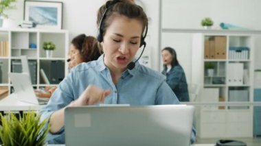 Neşeli Asyalı kadın çağrı merkezi uzmanı ofiste dizüstü bilgisayarla konuşuyor.