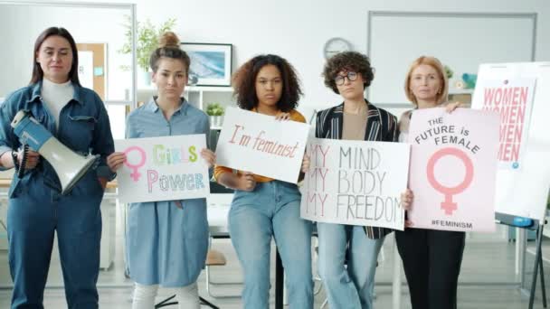 Sekelompok wanita dengan poster feminis berdiri di dalam ruangan dan melihat kamera dengan wajah serius — Stok Video