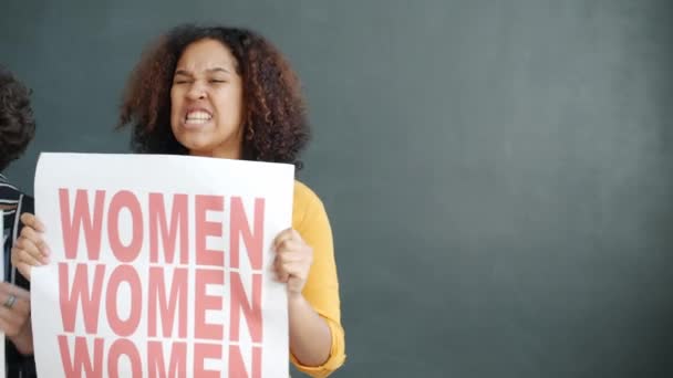 Gri arkaplana karşı durarak cinsiyet eşitliği çığlığı atan pankart tutan kadın aktivistlerin portresi — Stok video