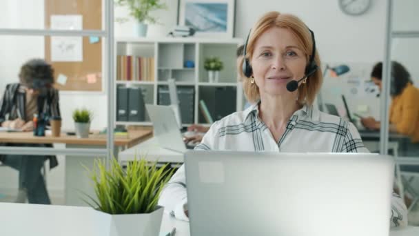 Retrato de especialista en call center señora madura sonriendo mirando a la cámara sentada en el lugar de trabajo — Vídeo de stock