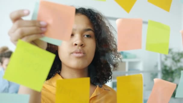 Detailní záběr seriózní afroamerické dívky, která si bere barevné samolepící poznámky ze skleněné tabule v kanceláři — Stock video