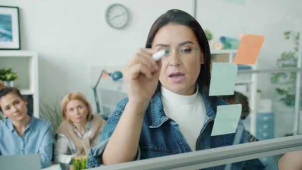 Seriös affärskvinna skriver på glasbräda med klibbiga anteckningar talar gör presentation för kollegor — Stockvideo