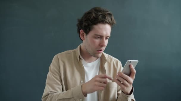 Wesoły młodzieniec wskazujący na ekran smartfona i uśmiechnięty polecający aplikację na szarym tle — Wideo stockowe