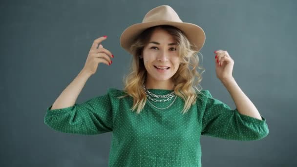 スタイリッシュな帽子をかぶって灰色の背景に笑みを浮かべて魅力的な若い女性の肖像画 — ストック動画