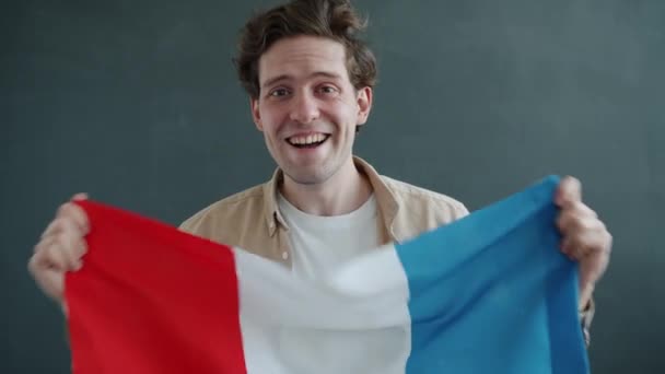 Fransız bayrağı tutan, gülümseyen ve gri arka plandaki kameraya bakan neşeli genç adamın yavaş çekim portresi. — Stok video