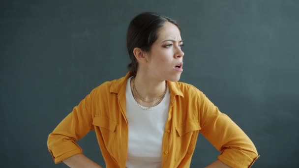 Портрет несчастной и злой молодой женщины, смотрящей в камеру, стоящую акимбо на сером фоне — стоковое видео