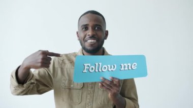 Afro-Amerikalı neşeli blogcu portresi Beni Takip Et tabelasına işaret ediyor ve beyaz arka planda gülümsüyor
