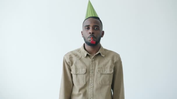Ritratto al rallentatore dell'uomo afroamericano che indossa un cappello luminoso che soffia il corno della festa con un volto miserabile — Video Stock