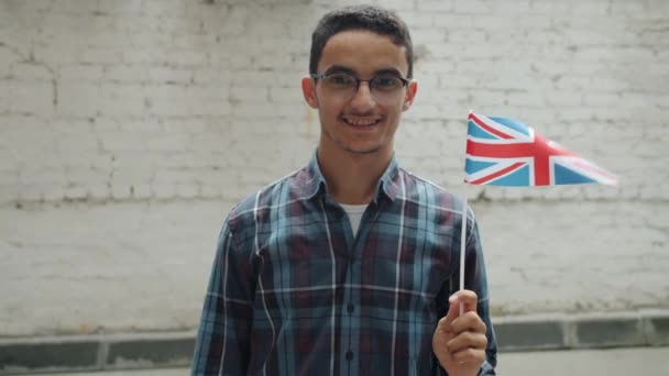 Slow motion of cheerful young Arab man waving British national flag and smiling standing in urban street — Stock Video