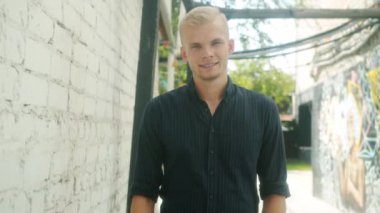 Yakışıklı genç adamın yavaş çekimde çekilmiş portresi. El sallıyor ve gülümsüyor. Şehir sokağının dışında duruyor.