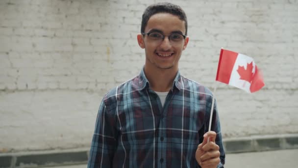 Movimento lento do homem do Oriente Médio acenando com a bandeira canadense e sorrindo de pé contra a parede de tijolos ao ar livre — Vídeo de Stock