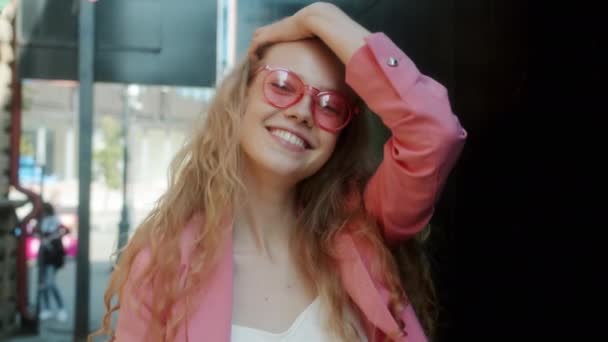 Повільний портрет милої дівчини в стильних рожевих сонцезахисних окулярах, що посміхаються стоячи зовні — стокове відео