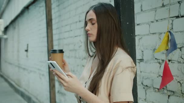 Töprengő fiatal nő megérinti okostelefon képernyő és az ivás, hogy menjen kávé szabadban a város utcájában — Stock videók