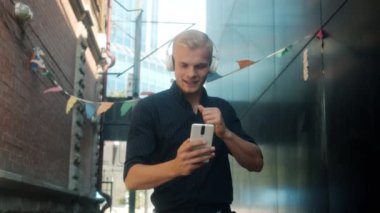Kulaklık takan yakışıklı sarışının ağır çekimde çekilmiş bir portresi. Elinde akıllı telefon, şehirde dans ediyor.