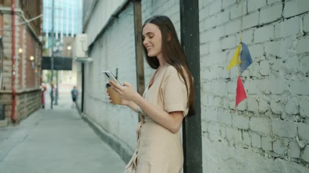 Powolny ruch radosnej pani za pomocą sdmartphone i śmiech trzymając filiżankę kawy na świeżym powietrzu w mieście — Wideo stockowe