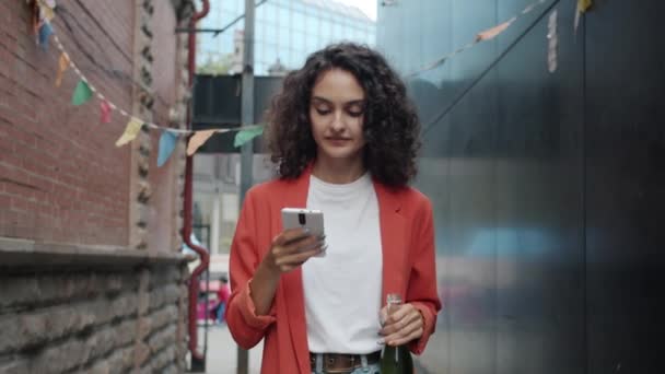 Akıllı telefon kullanan mutlu genç bir kadın yaz günü elinde içki şişesiyle şehirde yürüyor. — Stok video