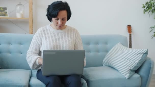 Зріла жінка використовує ноутбук, а потім насолоджується хорошими новинами, святкуючи успіх і посміхаючись вдома — стокове відео
