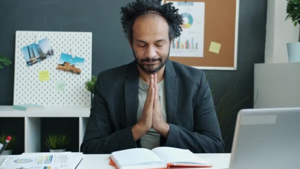 Pensativo hombre de Oriente Medio cogido de la mano en gesto namaste sentado en el escritorio con los ojos cerrados — Vídeo de stock