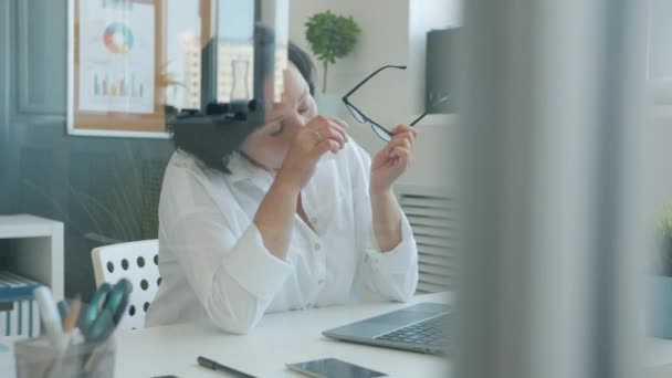 Уставшая деловая женщина снимает очки, страдающие от головной боли, работая с ноутбуком в офисе — стоковое видео