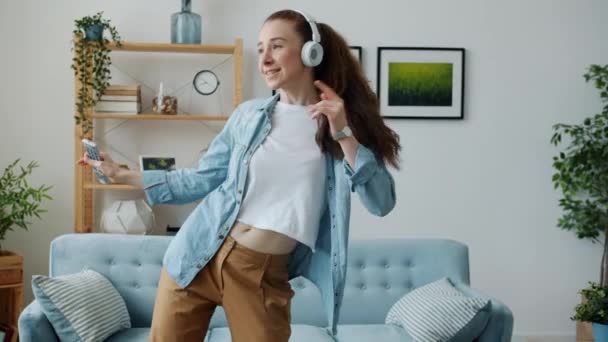 Estudiante juguetona cantando en control remoto y bailando escuchando música en auriculares en casa — Vídeo de stock