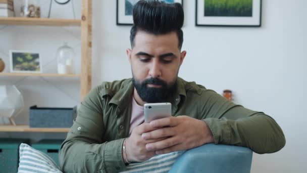 Elegante uomo di razza mista utilizzando smartphone che comunica sui social media sul divano di casa — Video Stock