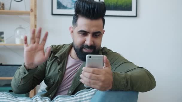 Χαρούμενος τύπος από τη Μέση Ανατολή που κάνει online βιντεοκλήση χαιρετώντας το χέρι και μιλώντας χρησιμοποιώντας το smartphone στο σπίτι — Αρχείο Βίντεο