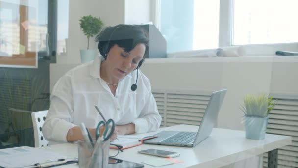 İş kadını kulaklık takıyor, yazı yazıyor ve dinliyor. Ofisteki laptop ekranına bakıyor. — Stok video