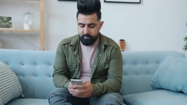 Moyen-Orient homme en utilisant smartphone écran tactile assis sur le canapé à la maison — Video