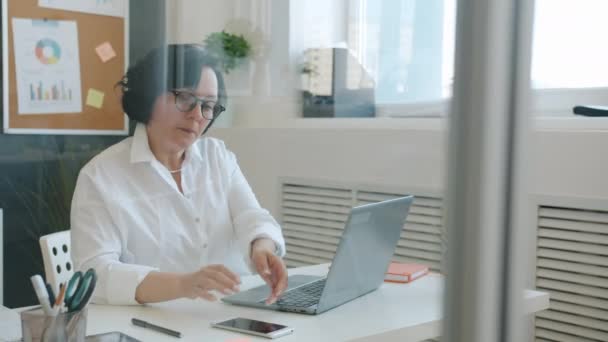 Mulher alegre usando laptop digitação, em seguida, falando no telefone celular e sorrindo trabalhando no escritório sozinho — Vídeo de Stock