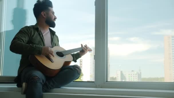 Baard gemengd ras gitarist zitten op vensterbank zingen gitaar spelen thuis — Stockvideo