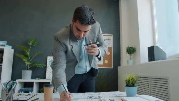 Empresário trabalhando com papéis e gravando mensagens no smartphone trabalhando no escritório sozinho — Vídeo de Stock