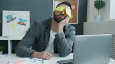 İş yerinde masa başında uyuyan beceriksiz bir ofis çalışanı. Yüzünde açık gözler gösteren yapışkan notlarla.