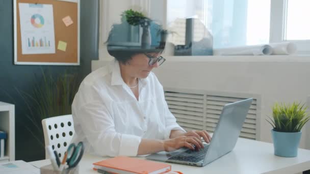 Медленное движение зрелой женщины, работающей с компьютером ноутбука, печатающей сидя за столом в офисе — стоковое видео