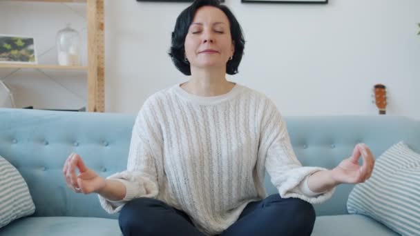 Señora madura sentada en sofá en pose de loto y meditando con los ojos cerrados en casa — Vídeos de Stock