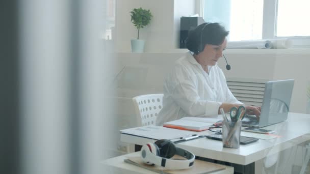 Movimento lento de empresária usando fones de ouvido digitando com laptop e escrevendo no escritório — Vídeo de Stock